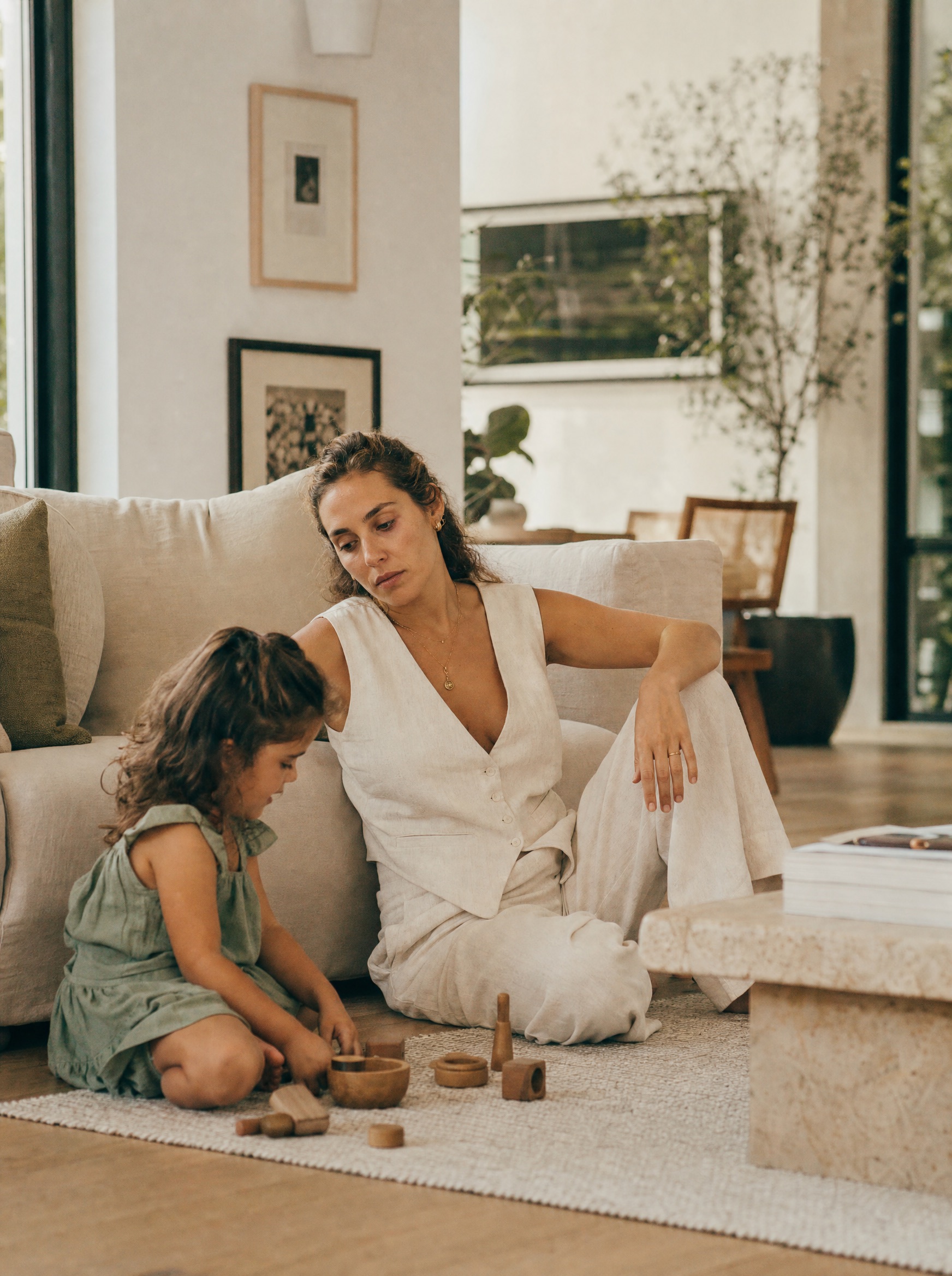 Mamá sentada en el piso recargada en el sillón, acompañando a su hija que juega en el tapete, con la mirada cansada de una tarde larga de agosto