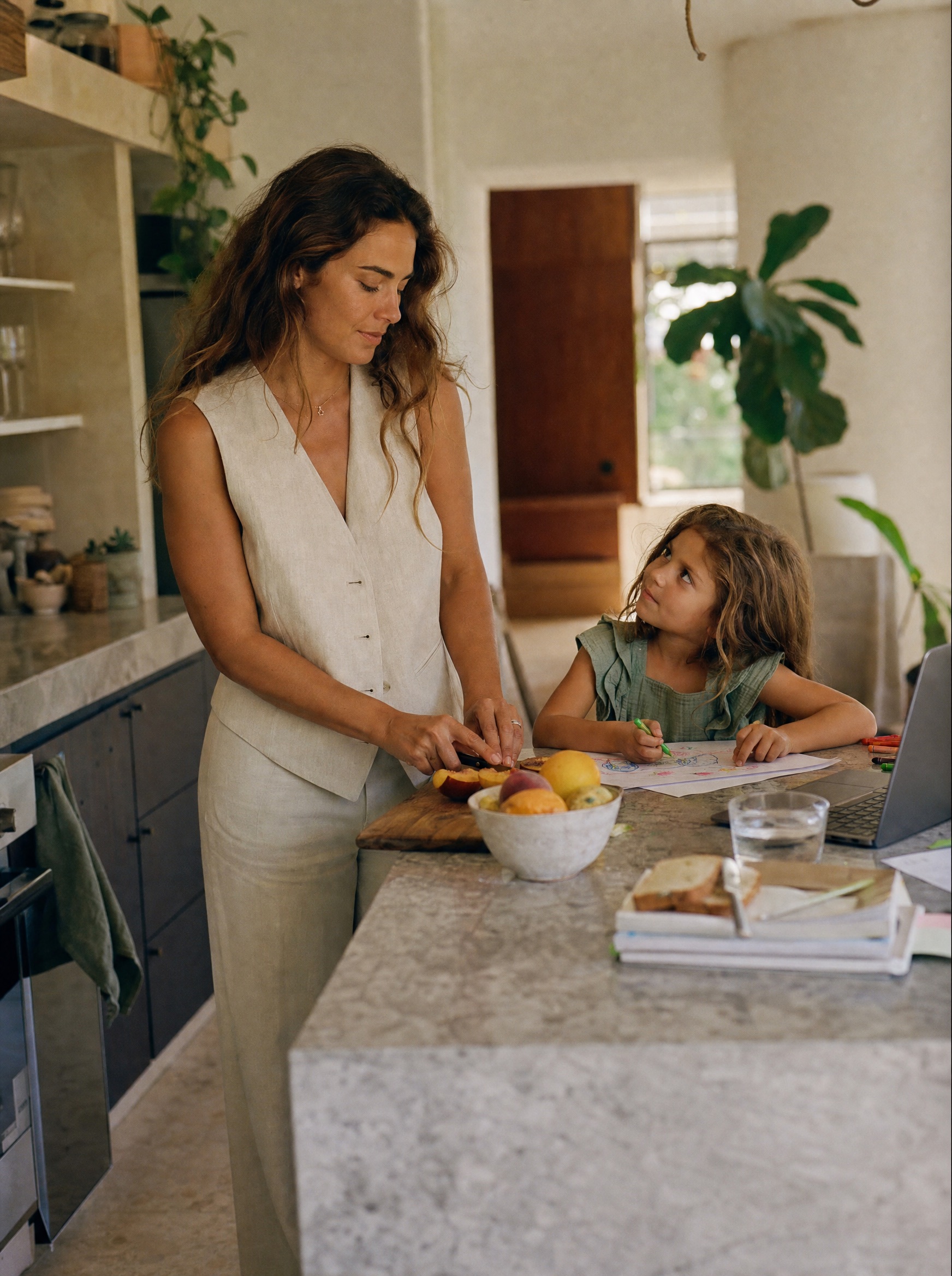 Mamá preparando la comida en la cocina mientras su hija dibuja en la barra, con la laptop abierta a un lado
